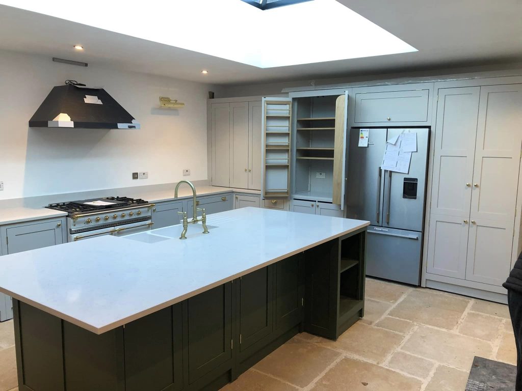 A finished kitchen with pale grey shaker units, a dark green island topped in white quartz, brass tapware, a range cooker and a roof lantern above flagstone flooring.