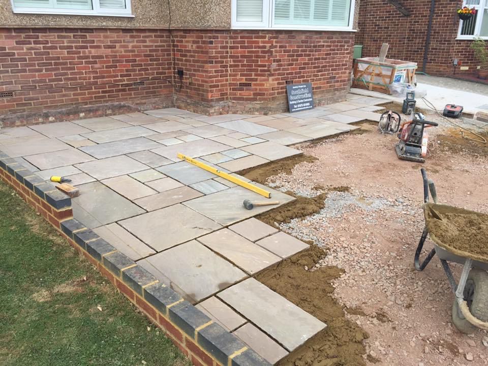 Indian sandstone patio being laid against a red brick house, edged with a charcoal brick course, with a spirit level checking the falls and a Southfield Construction site board behind.