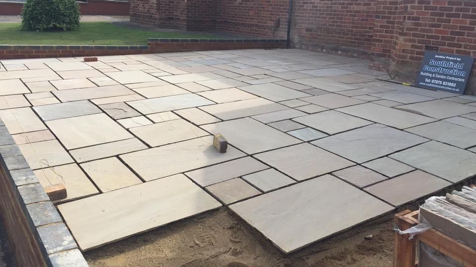 A large Indian sandstone patio laid across a rear garden against a red brick house, edged with a charcoal brick course, with a Southfield Construction site board standing in the corner.