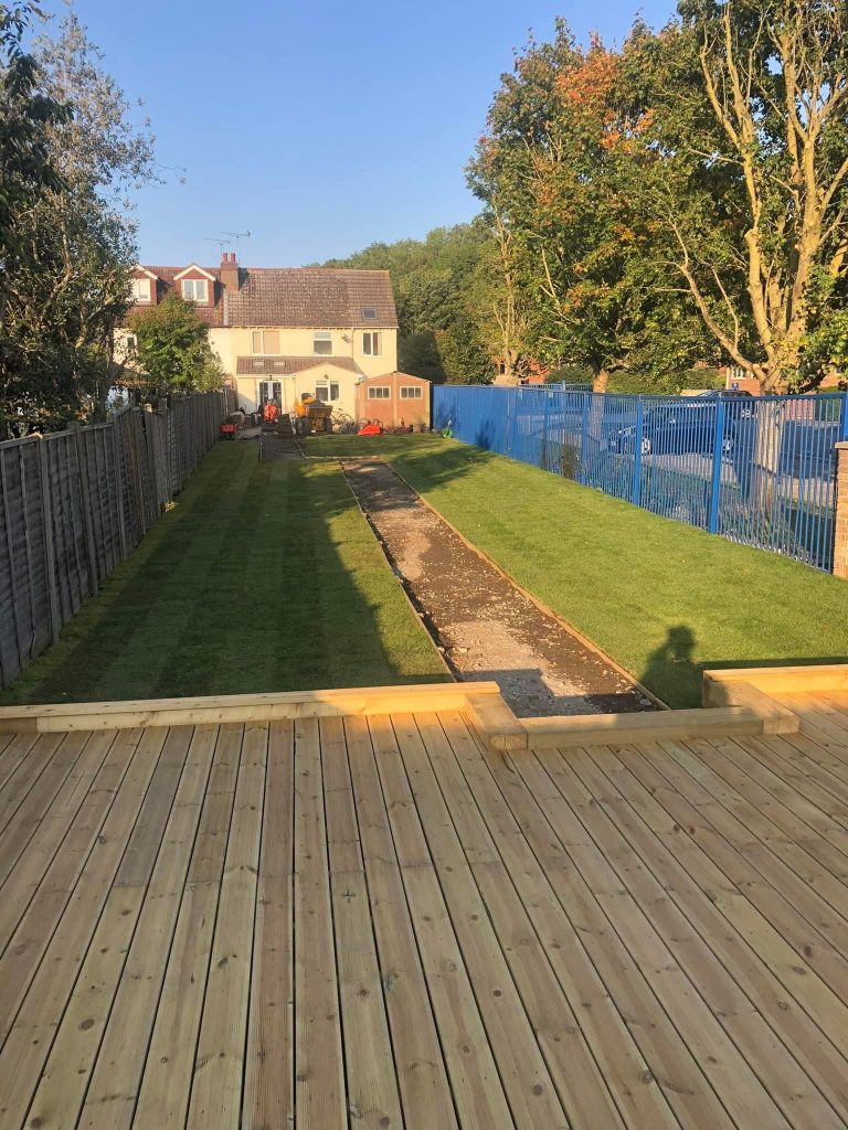 New timber decking in the foreground looking down a long garden of freshly laid striped turf towards a rendered semi-detached house, with a tracked dumper parked by the back door.