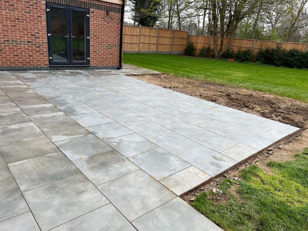 Grey porcelain paving with spacers still in place, running alongside a red brick wall with anthracite French doors, beside a newly turfed lawn and close board fencing.