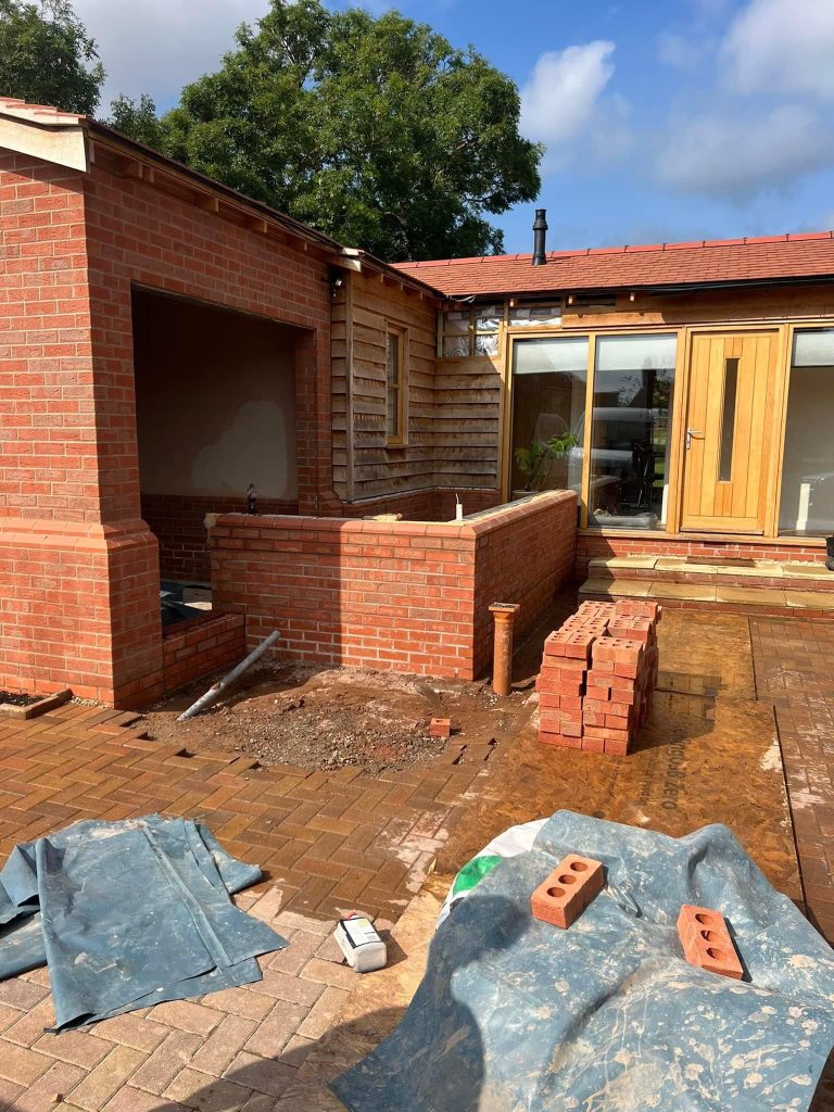 A single storey brick and cedar clad extension under construction, with a new brick garden wall, oak framed patio doors and a clay tiled roof, and bricks stacked on the block paving ready for the next lift.